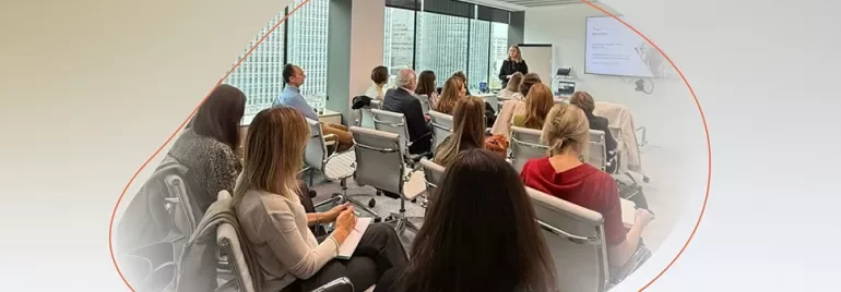 Group attending a presentation in a modern meeting room, with a speaker standing at the front near a screen.